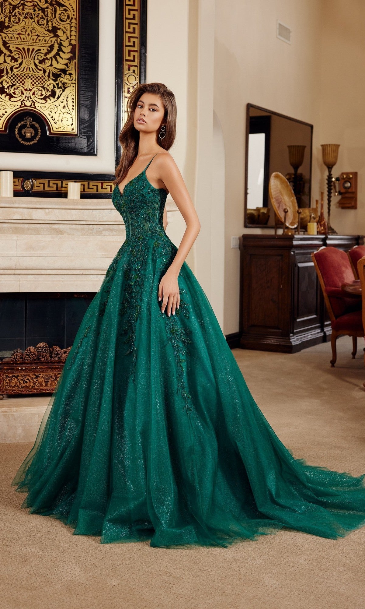 A woman stands indoors wearing the Nox Anabel H1883 long prom dress in emerald, featuring a fitted corset bodice, flared skirt, sparkling details, embroidery, and delicate floral appliques.